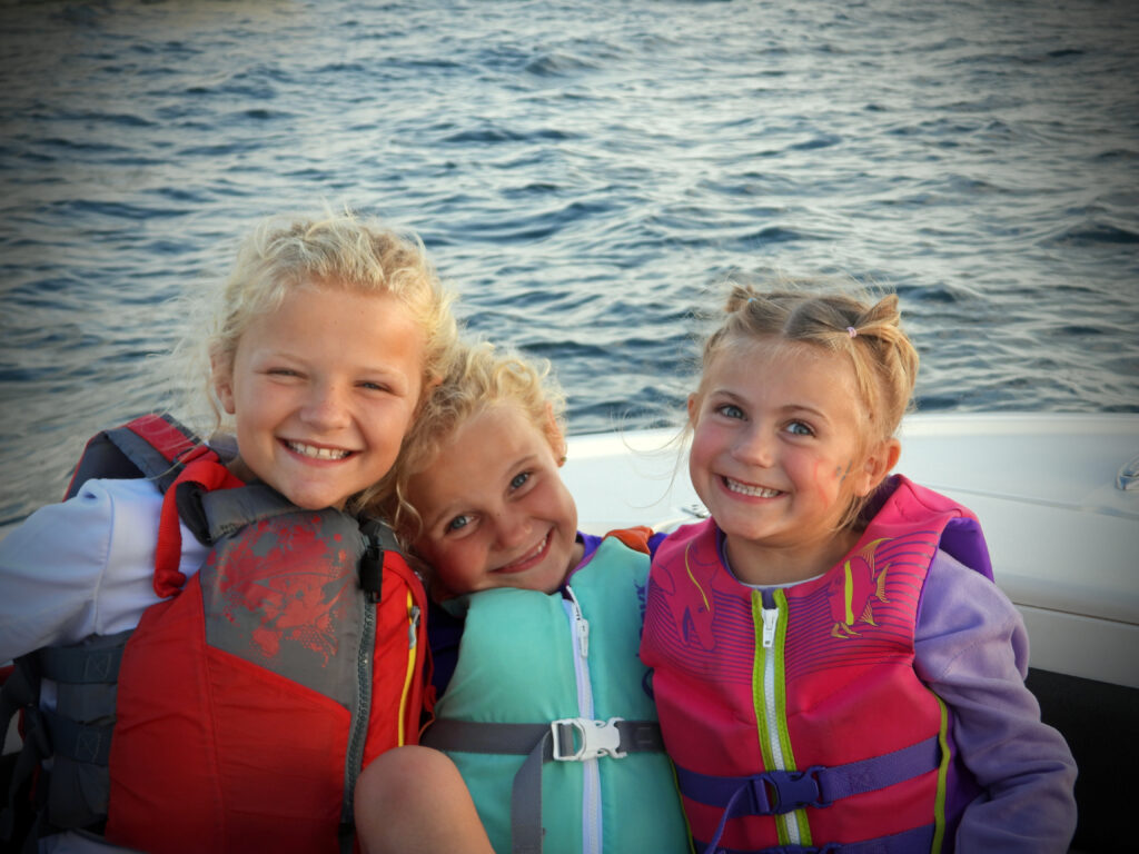 Three smiling children wearing colorful life jackets sitting together on a boat with water in the background