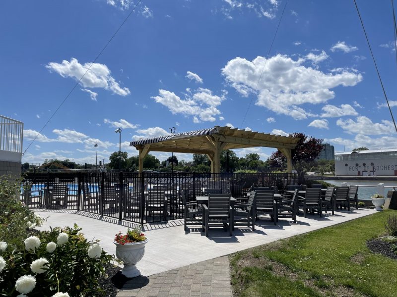 Outdoor patio area with multiple dark wooden chairs arranged around small tables on a concrete surface. The seating area is partially shaded by a wooden pergola with a striped canopy. The patio is enclosed by a black metal fence and surrounded by green grass and flower beds with white and red flowers. In the background, there is a river or canal with a large white building labeled ‘Port of Rochester.’ The sky is bright blue with scattered white clouds