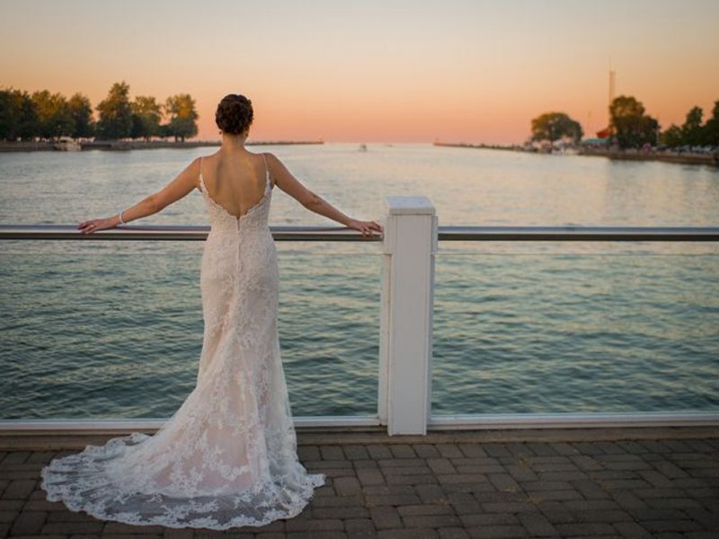 Bride in a lace wedding gown standing with her arms resting on a railing overlooking water at sunset.