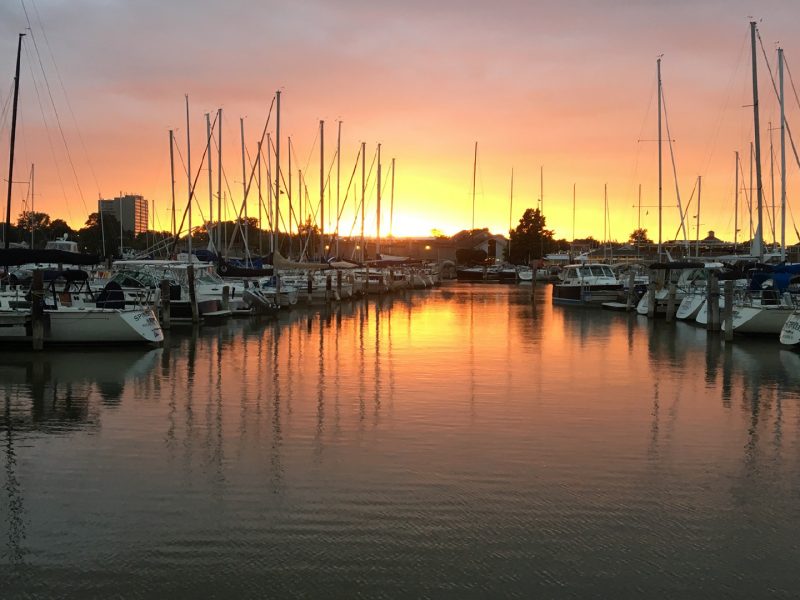 Boats docked in a marina at sunset with orange and pink sky reflecting on calm water.
